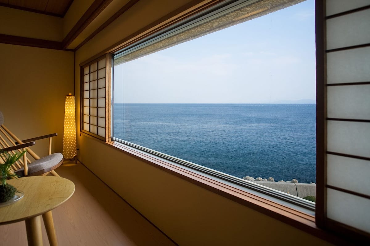 Ein Zimmer mit Meerblick, ein Holzstuhl, ein kleiner Tisch mit einer Pflanze und eine traditionelle japanische Laterne.
