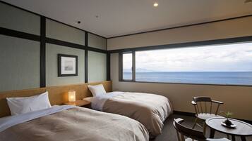 A hotel room with two beds, a small table, a chair, and a large window with a view of the sea.