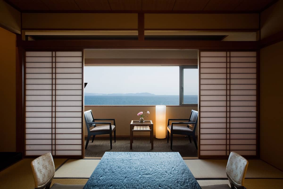 Ein traditioneller japanischer Raum mit Tatami-Böden, einem niedrigen Tisch mit Blumenvase, zwei Stühlen und Meerblick durch ein großes Fenster.