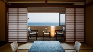 Ein traditioneller japanischer Raum mit Tatami-Böden, einem niedrigen Tisch mit Blumenvase, zwei Stühlen und Meerblick durch ein großes Fenster.