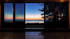 Ein Zimmer mit großen Fenstern, das Blick auf das Meer und Bäume im Abenddämmerung bietet.