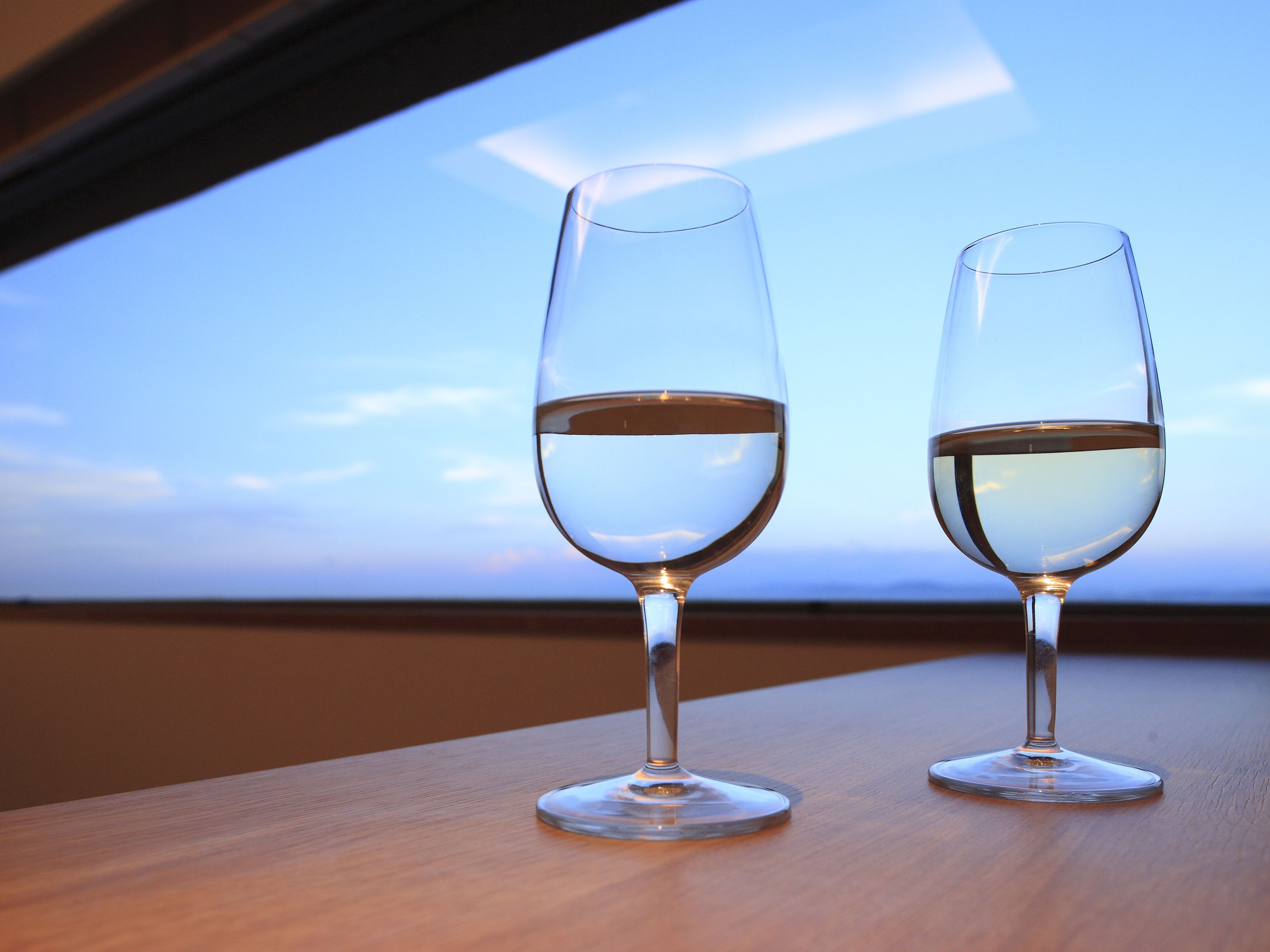 Two wine glasses on a wooden table with a view of the sky through a window.