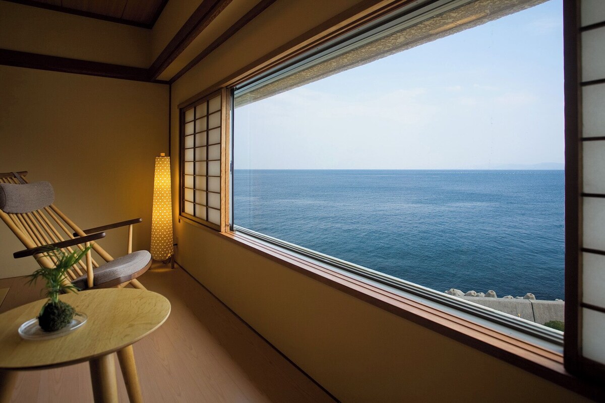 Una habitación con vista al mar, un futón de tatami, una mesa baja con una planta y una lámpara tradicional japonesa.