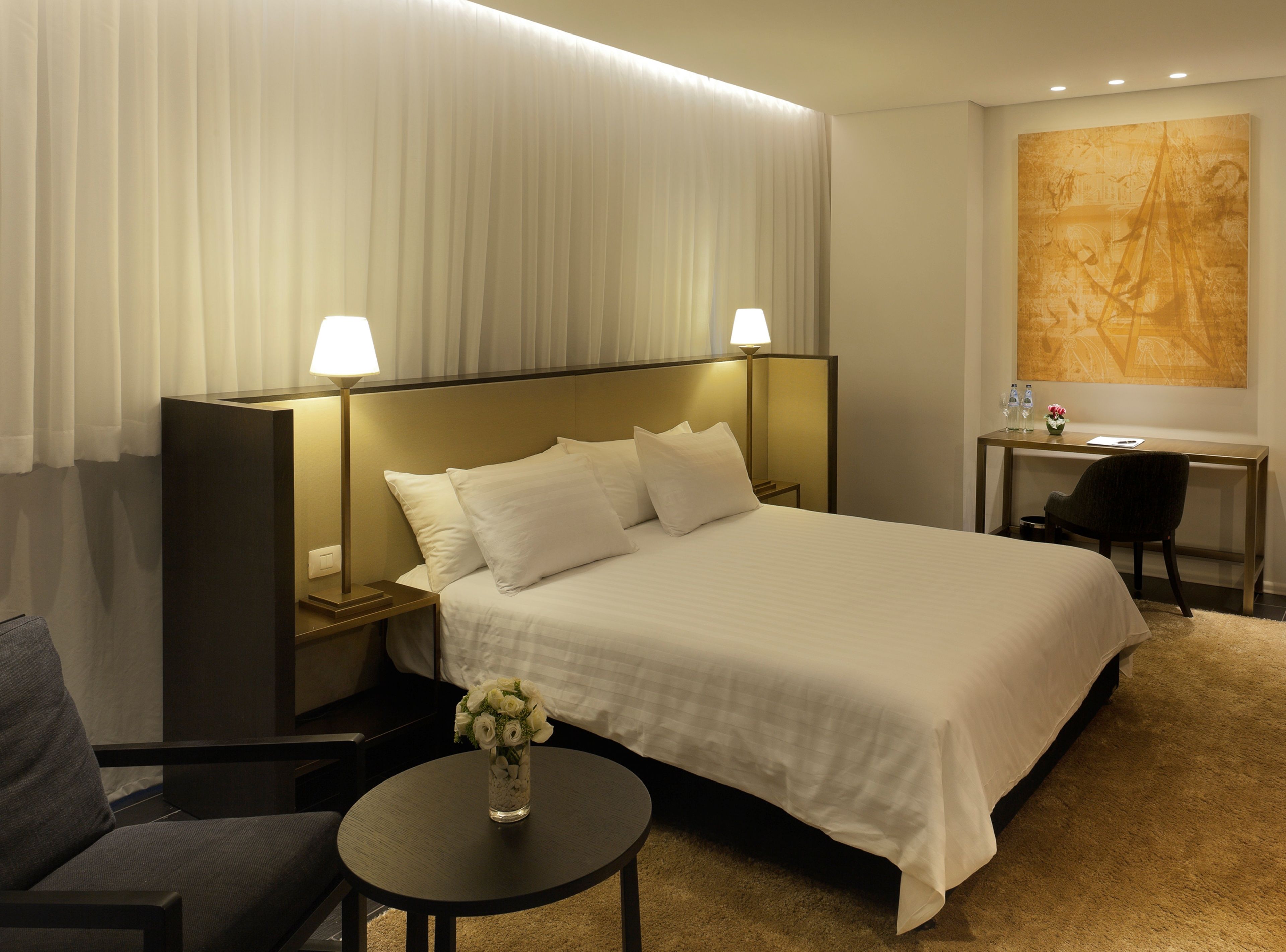 Een hotelkamer met een groot bed, twee bedlampen, een bureau met stoel, een klein tafeltje met een bloevas, en een groot abstract schilderij aan de muur.