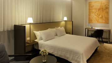Een hotelkamer met een groot bed, twee bedlampen, een bureau met stoel, een klein tafeltje met een bloevas, en een groot abstract schilderij aan de muur.