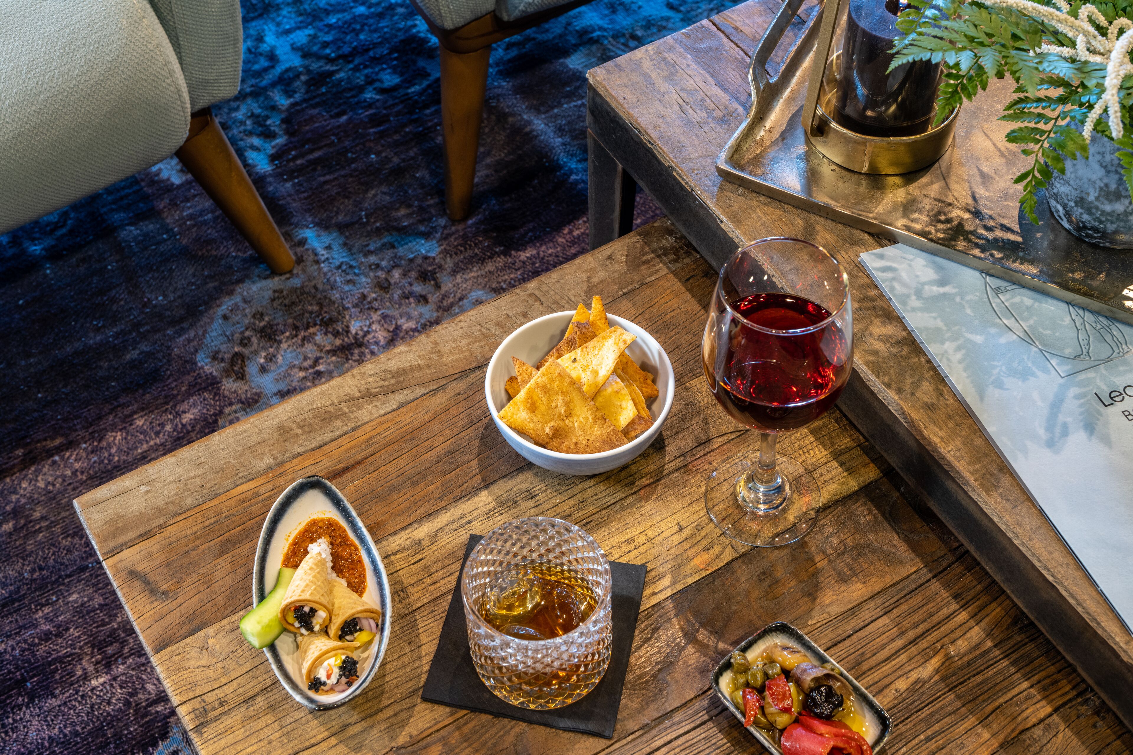 Ein Holztisch mit einer Schüssel Chips, einem Glas roter Flüssigkeit und einer kleinen Olivenplatte.
