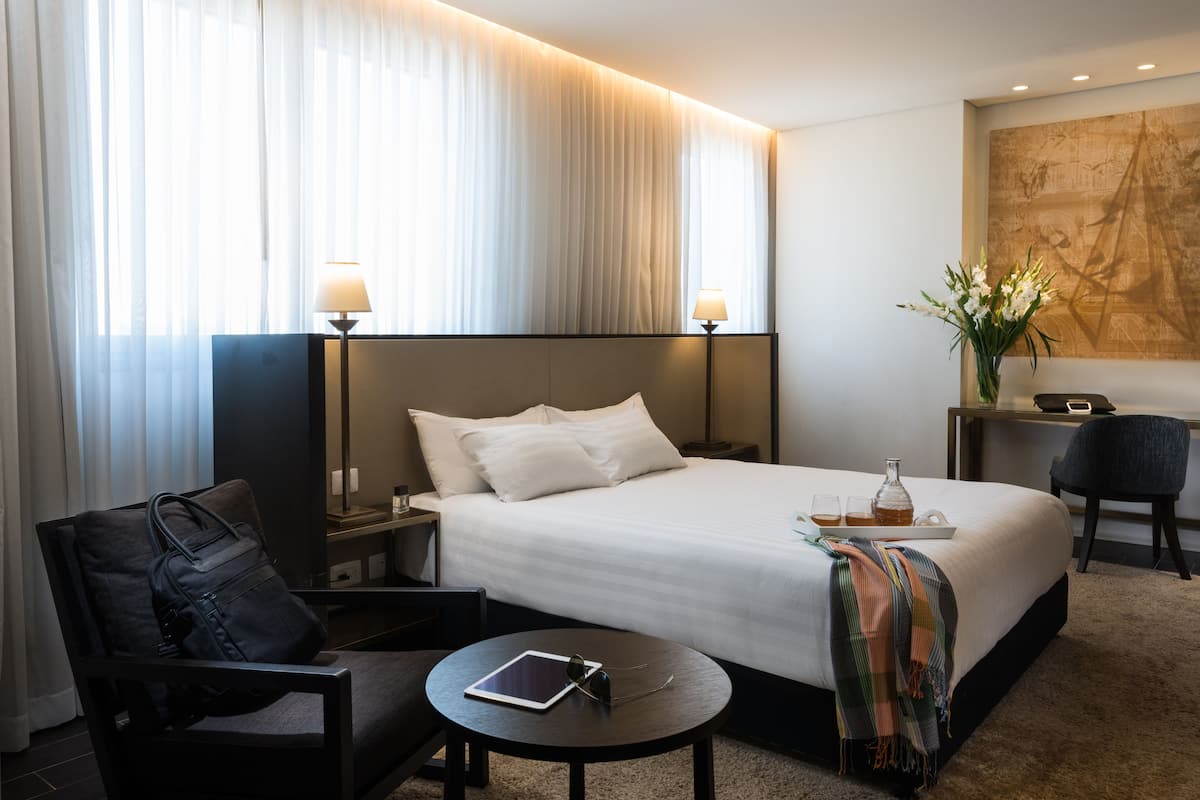 A hotel room with a large bed, a bedside table with a lamp, a chair, a small round table with a tablet and sunglasses, and a vase with flowers.