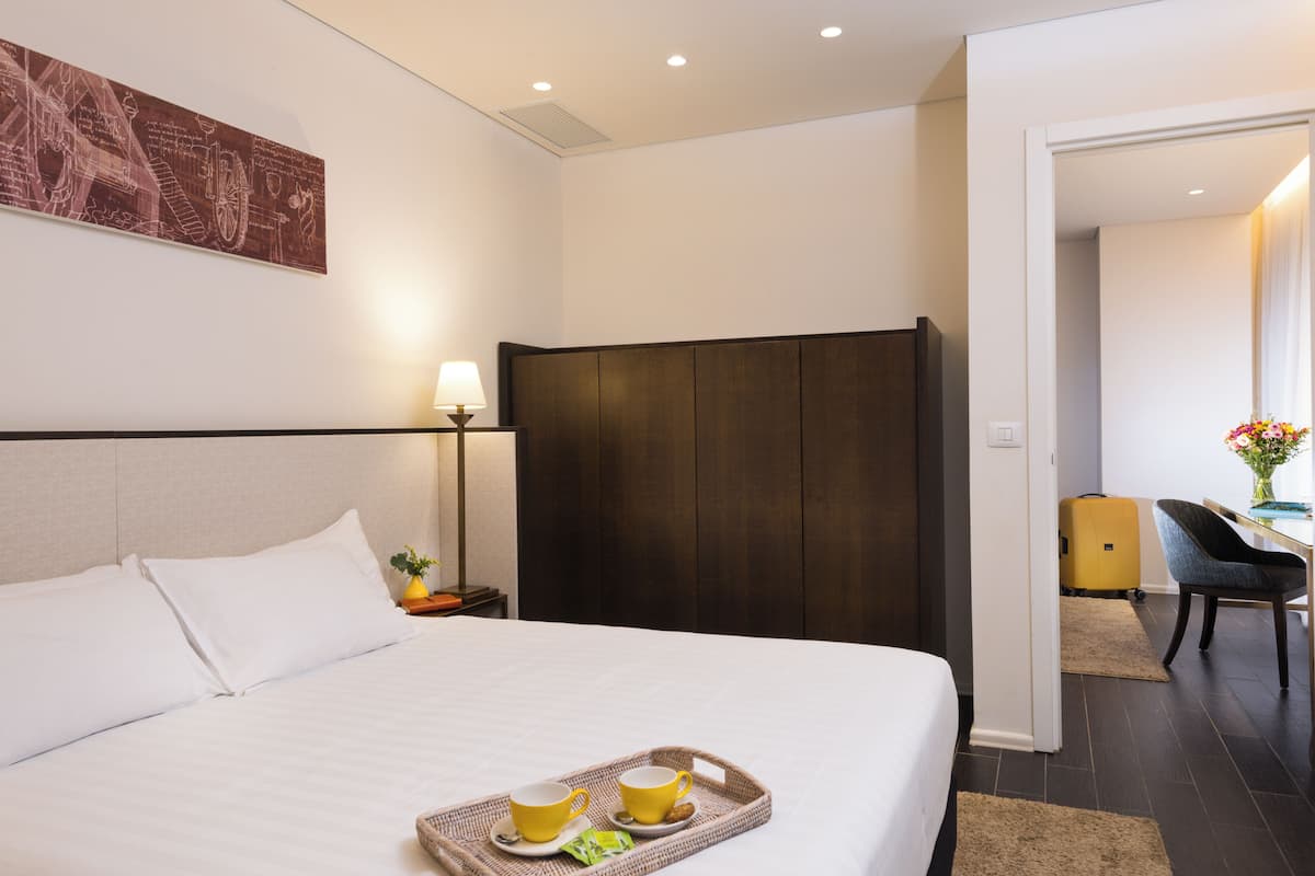 A hotel room with a large bed, a nightstand with a lamp, a wooden wardrobe, a desk with a chair, and a small table with a vase of flowers.