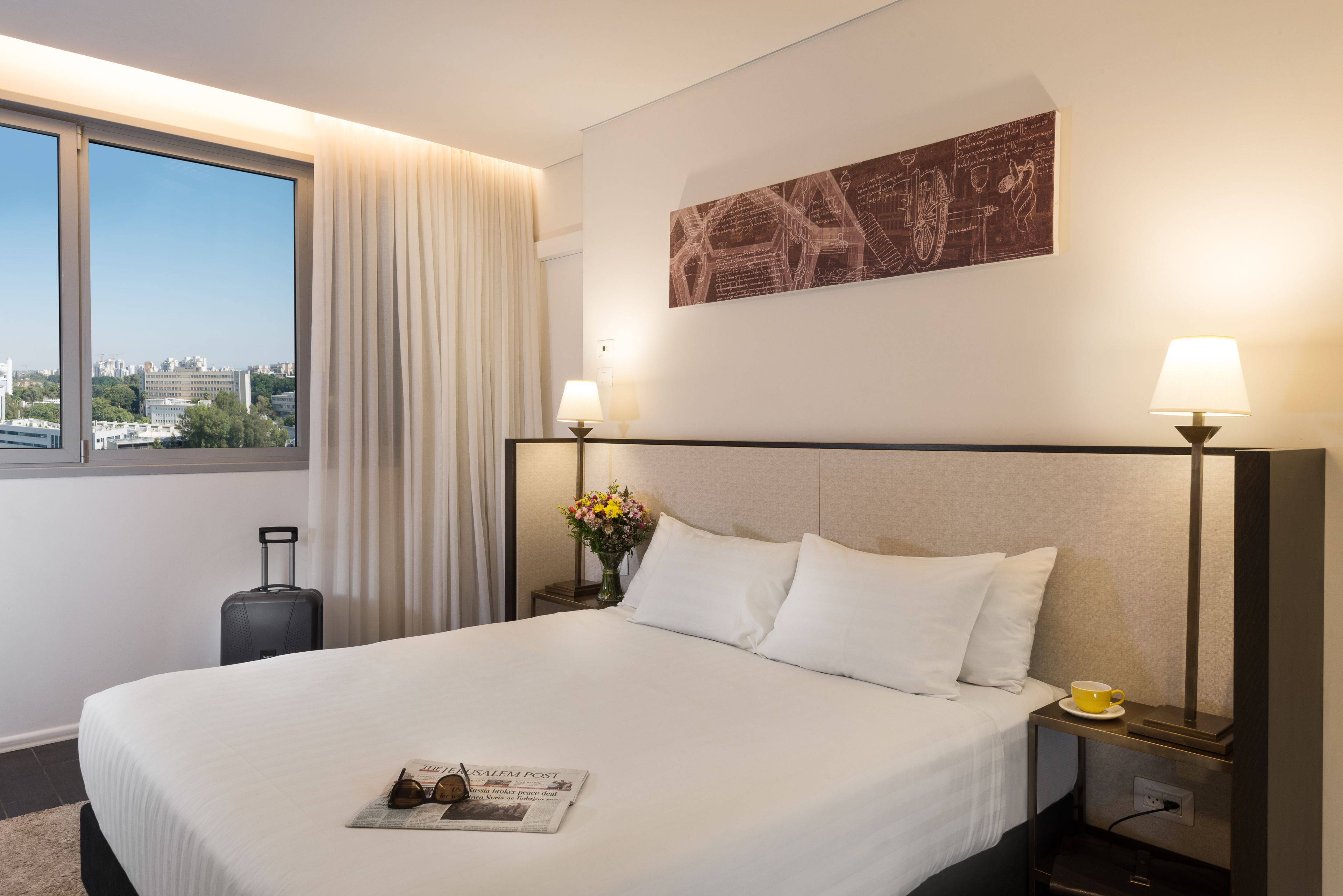 Habitación de hotel con una cama grande, vistas a la ciudad, una ventana con cortinas, una mesita de noche con lámpara y una maleta.