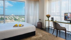 A hotel room with a large bed, a desk with a chair, a TV, and a view of the city.