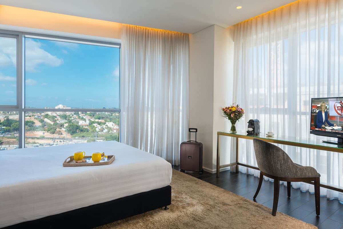 A hotel room with a large bed, a desk with a chair, a TV, and a view of the city.