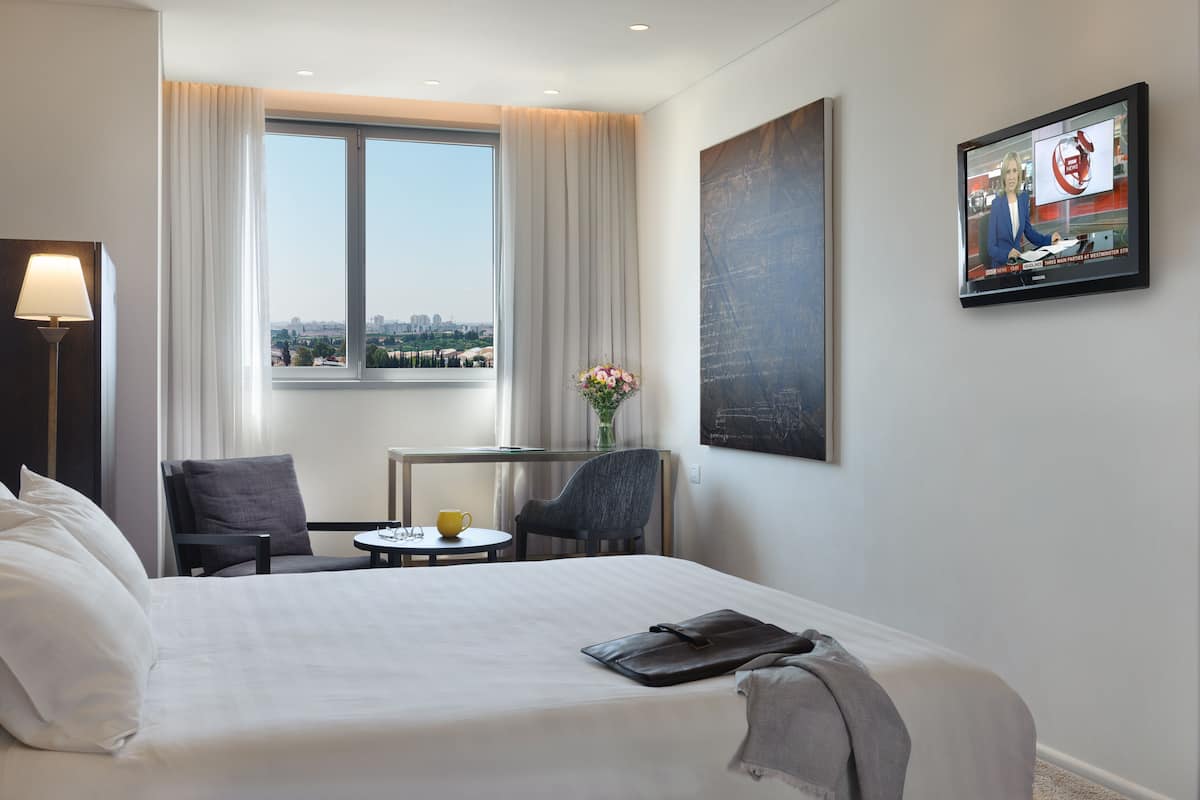A hotel room with a large bed, a desk with a chair, a small table with a cup, and a window with curtains.
