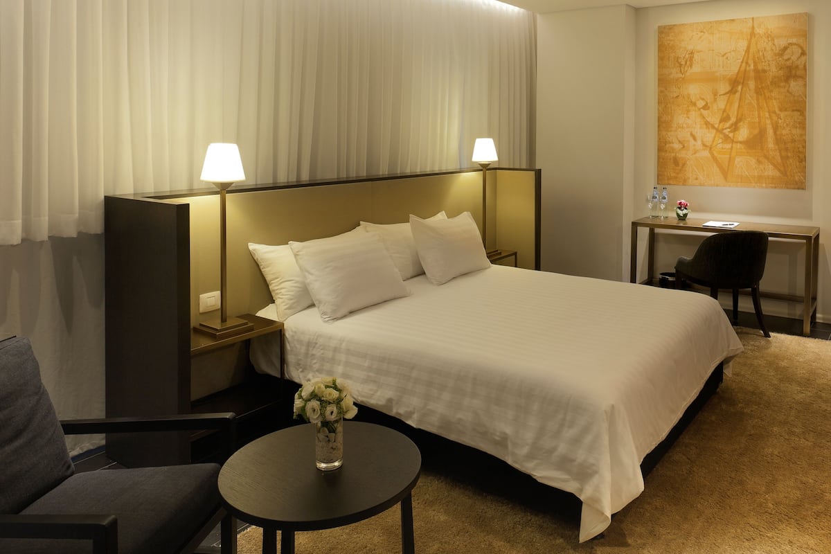 A hotel room with a large bed, bedside lamps, a desk with a chair, a small table with a vase of flowers, and a large abstract painting on the wall.