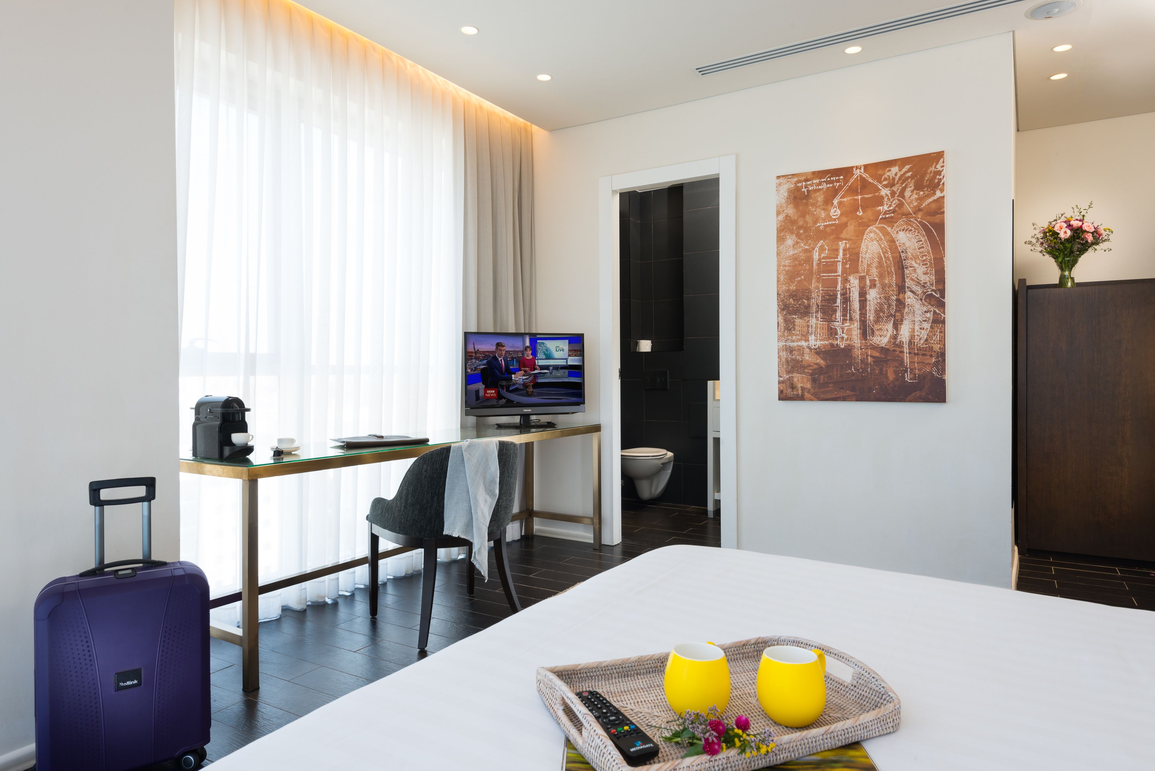 Habitación de hotel con cama, escritorio, televisor, maleta y bandeja con dos tazas y un control remoto.