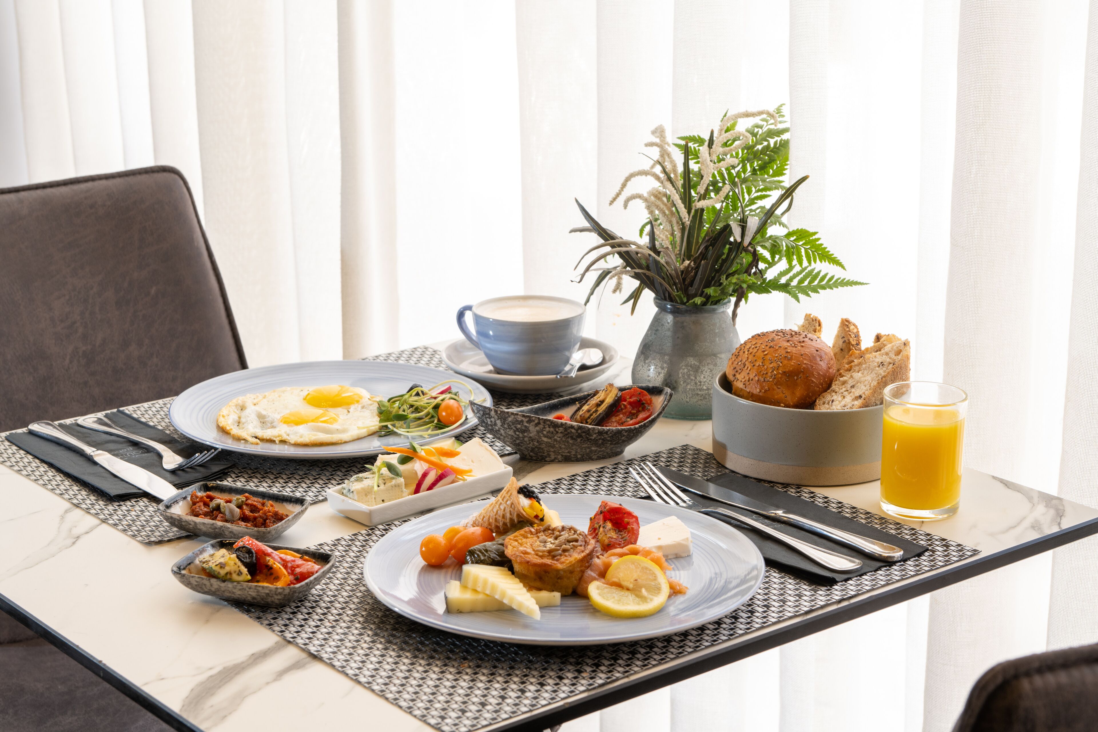 Una mesa de desayuno con platos de comida, una taza de café, un vaso de jugo de naranja y un florero con plantas.