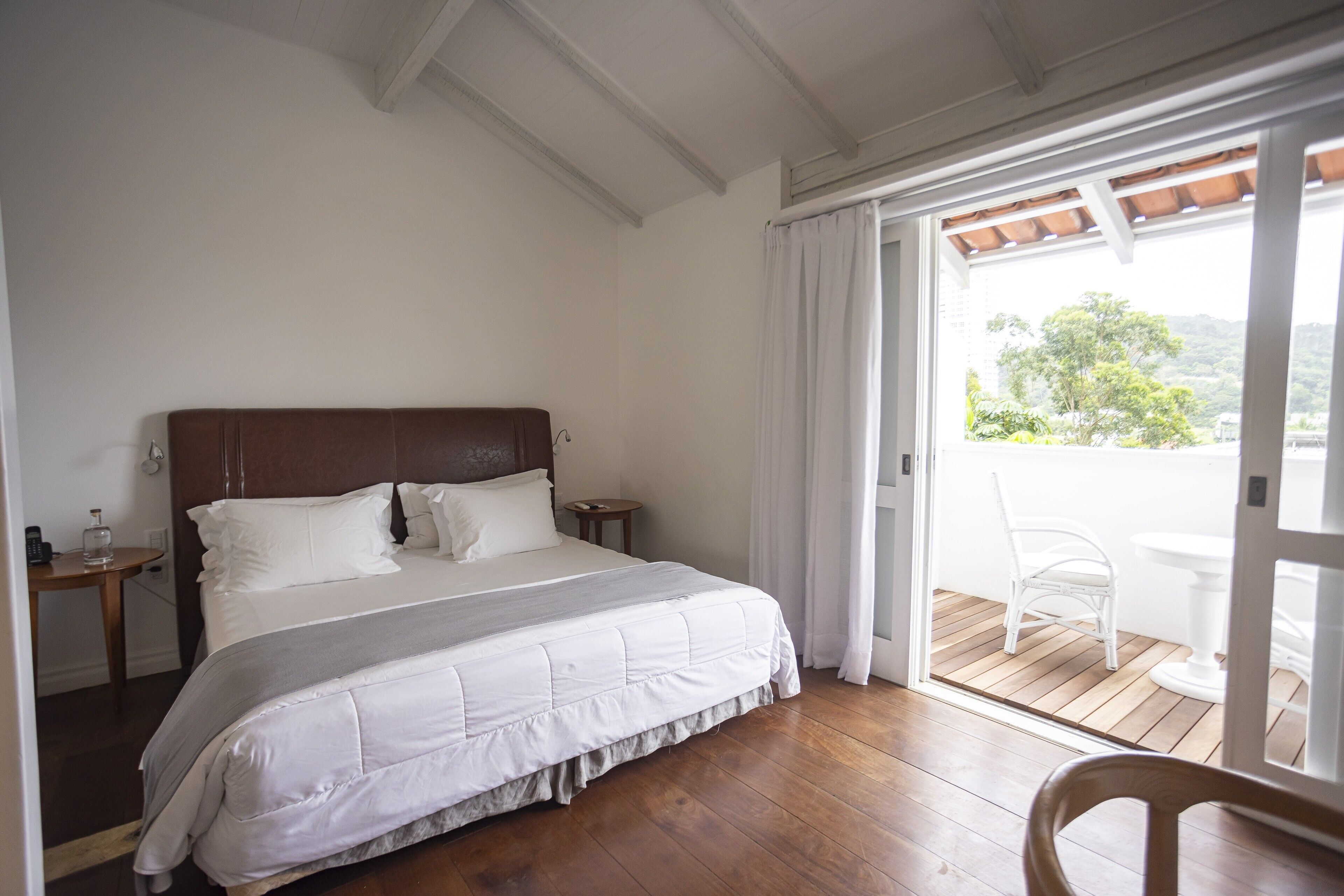 Un dormitorio con una cama grande, suelo de madera, un balcón con una silla y vistas a zonas verdes.