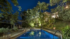 Une piscine entourée d’une végétation luxuriante et dotée d’une terrasse en bois, avec un bâtiment visible au loin.
