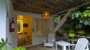 Une terrasse en bois avec des chaises blanches, une lampe suspendue et une porte donnant sur une pièce où se trouvent un lit et un téléviseur.