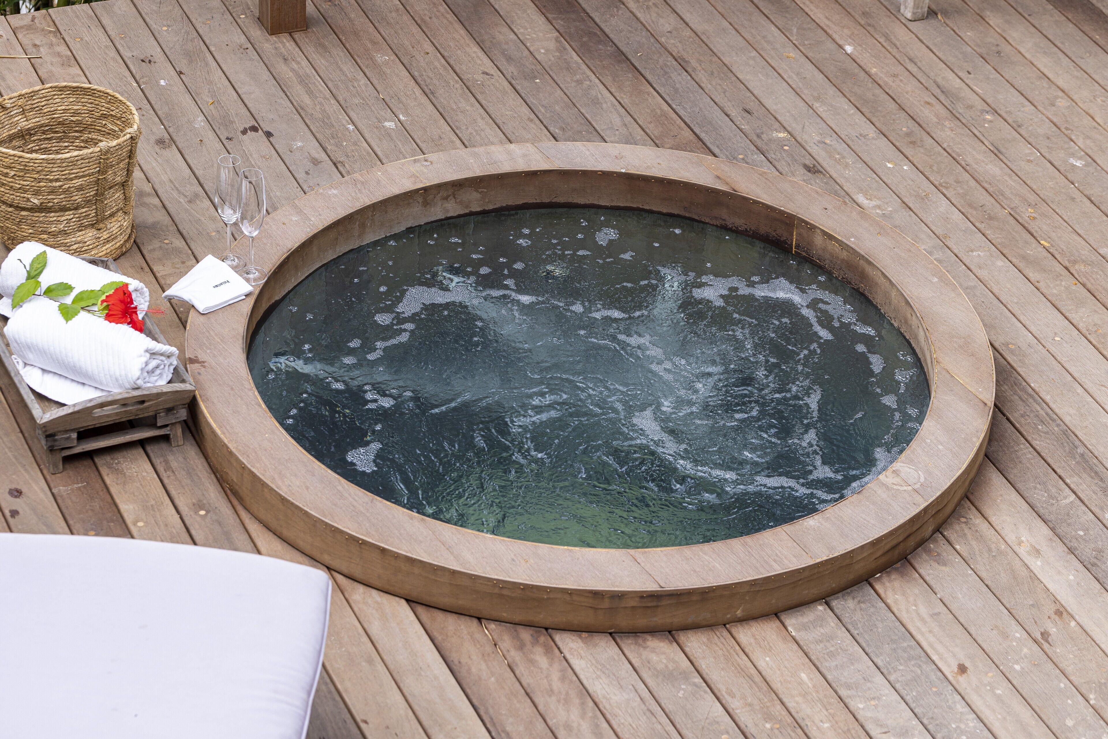 A wooden deck with a circular hot tub, a wicker basket, and a white towel with a red flower.