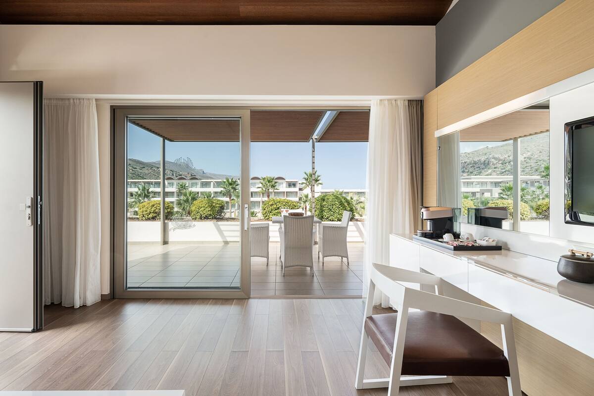A modern hotel room with a sliding glass door leading to a balcony with a view of other hotel buildings and palm trees.
