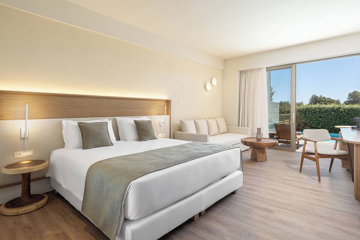 Habitación de hotel moderna con una cama grande, un sofá, un comedor y vistas al exterior.
