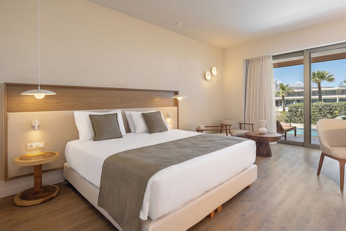 A hotel room with a large bed, a wooden headboard, a round wooden table, a chair, and a view of a pool and palm trees.