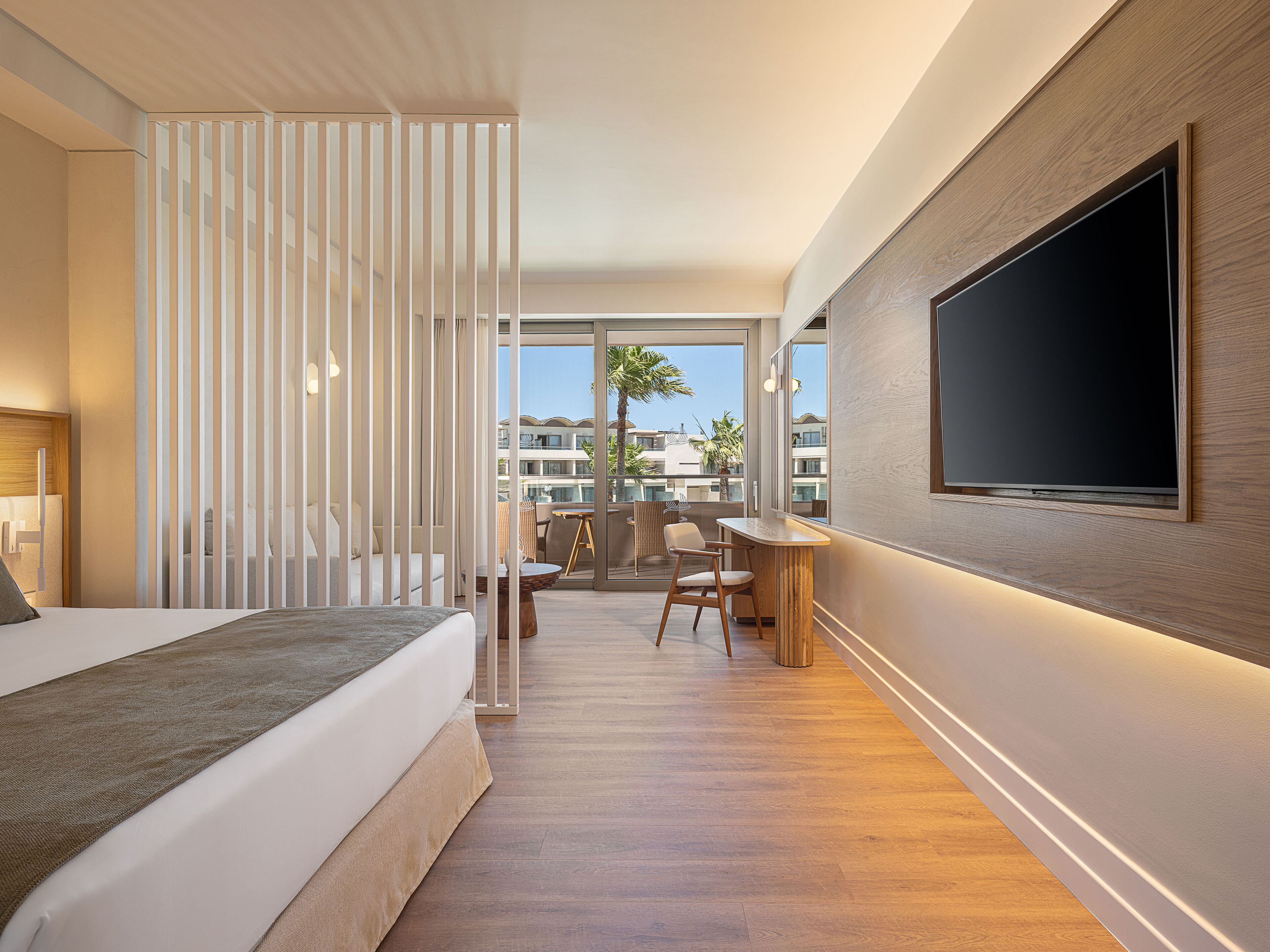 Una habitación de hotel moderna con una cama grande, un escritorio con silla, un televisor montado en la pared y un balcón con vistas a otros edificios.