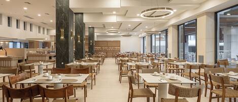 Un amplio comedor de hotel con numerosas mesas y sillas, ventanas grandes y modernas lámparas.