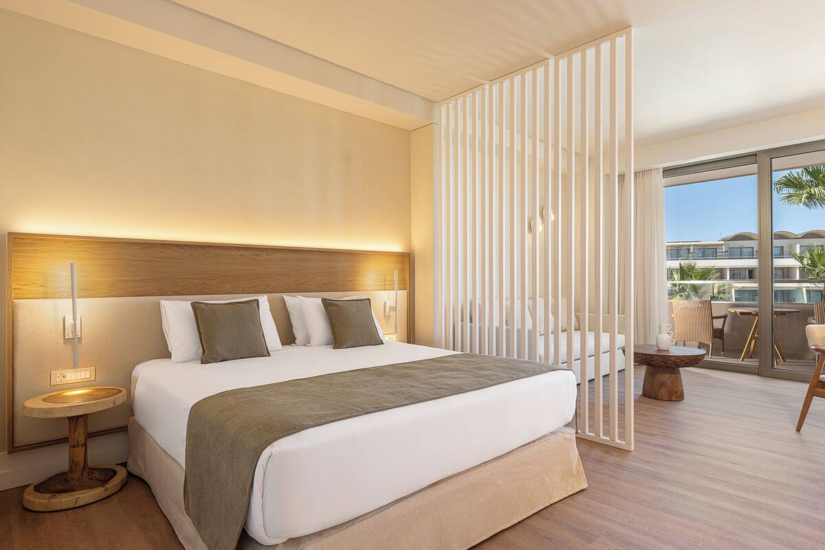 A modern hotel room with a large bed, a bedside table, a small round table, and a view of the pool and buildings outside.