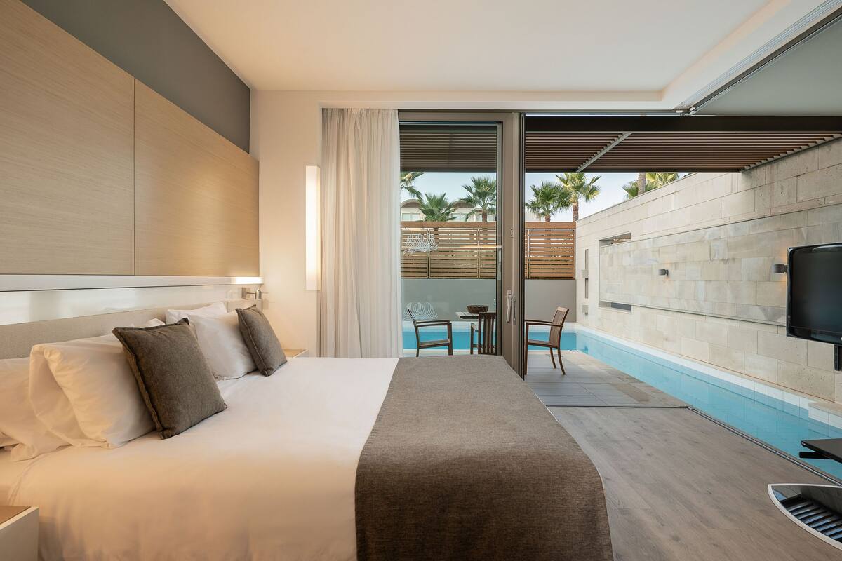 Ein Hotelzimmer mit einem großen Bett, einem an der Wand befestigten Fernseher, einer kleinen Poolanlage mit Tisch und Stühlen sowie Blick auf Palmen und ein Gebäude im Hintergrund.