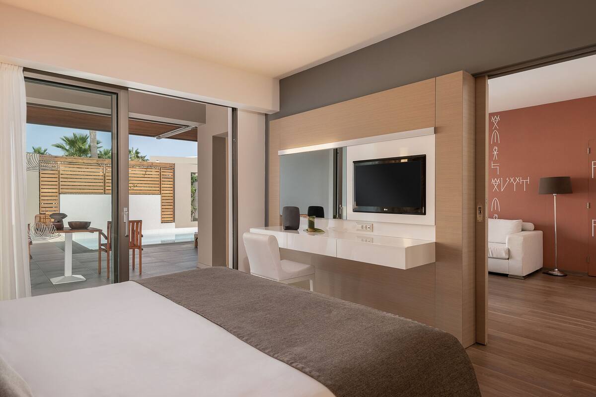 Habitación de hotel moderna con una cama grande, un escritorio con televisor de pantalla plana, un sofá blanco y un ventanal corredero que da a un área exterior con mesa y sillas.