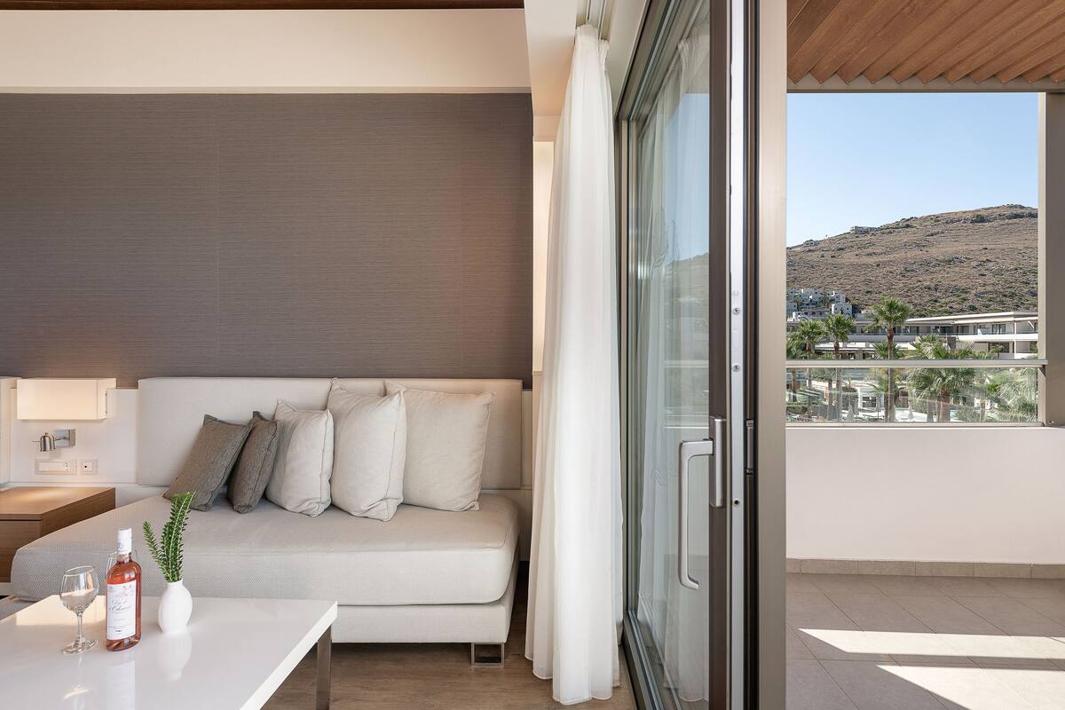 Una habitación moderna con sofá, una mesa con botella y vaso, y vistas a una ladera a través de ventanas amplias.