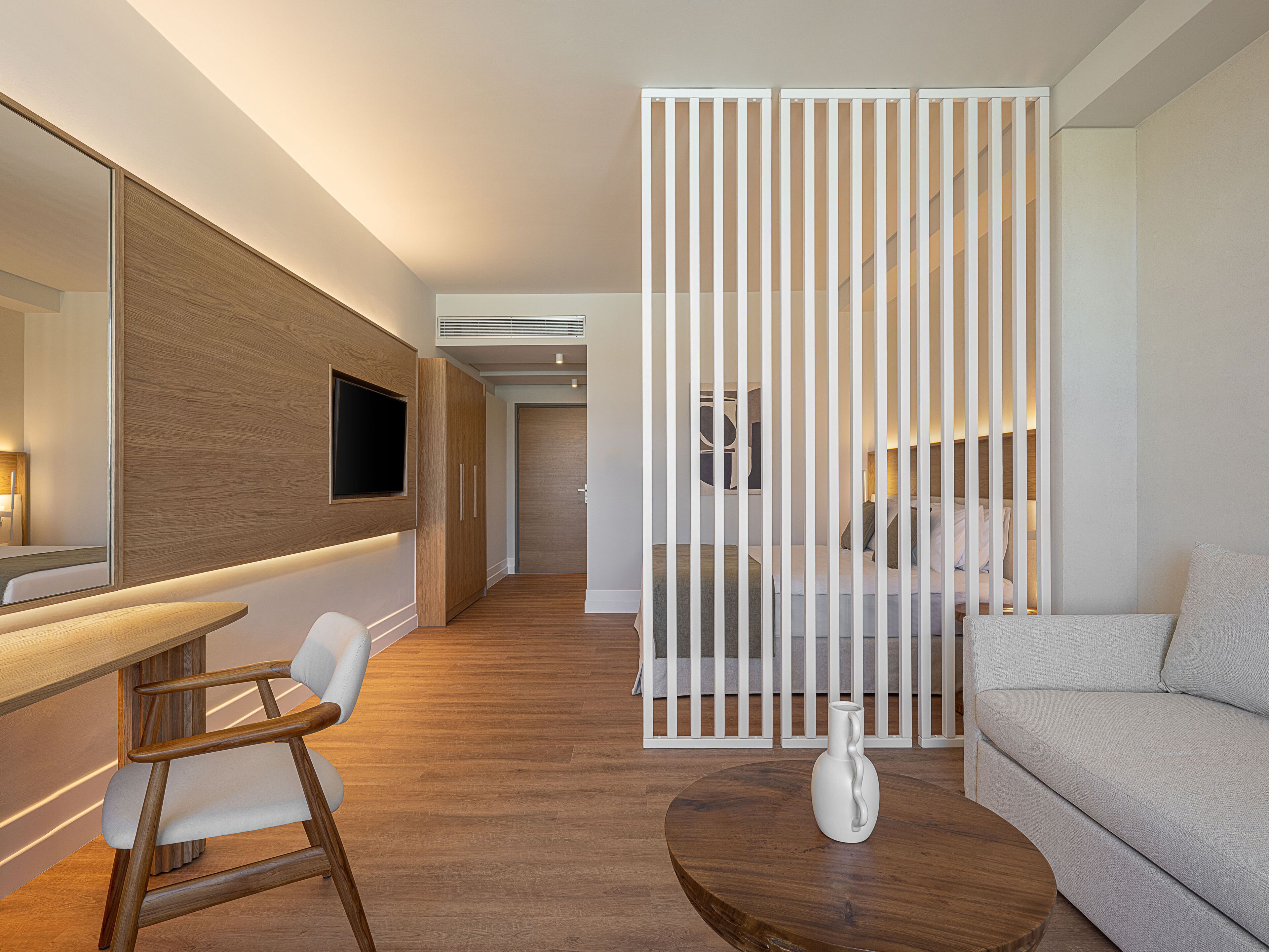 Una habitación de hotel moderna con un escritorio de madera, una silla blanca, una mesa redonda de centro de madera, un sofá blanco y un jarrón decorativo.