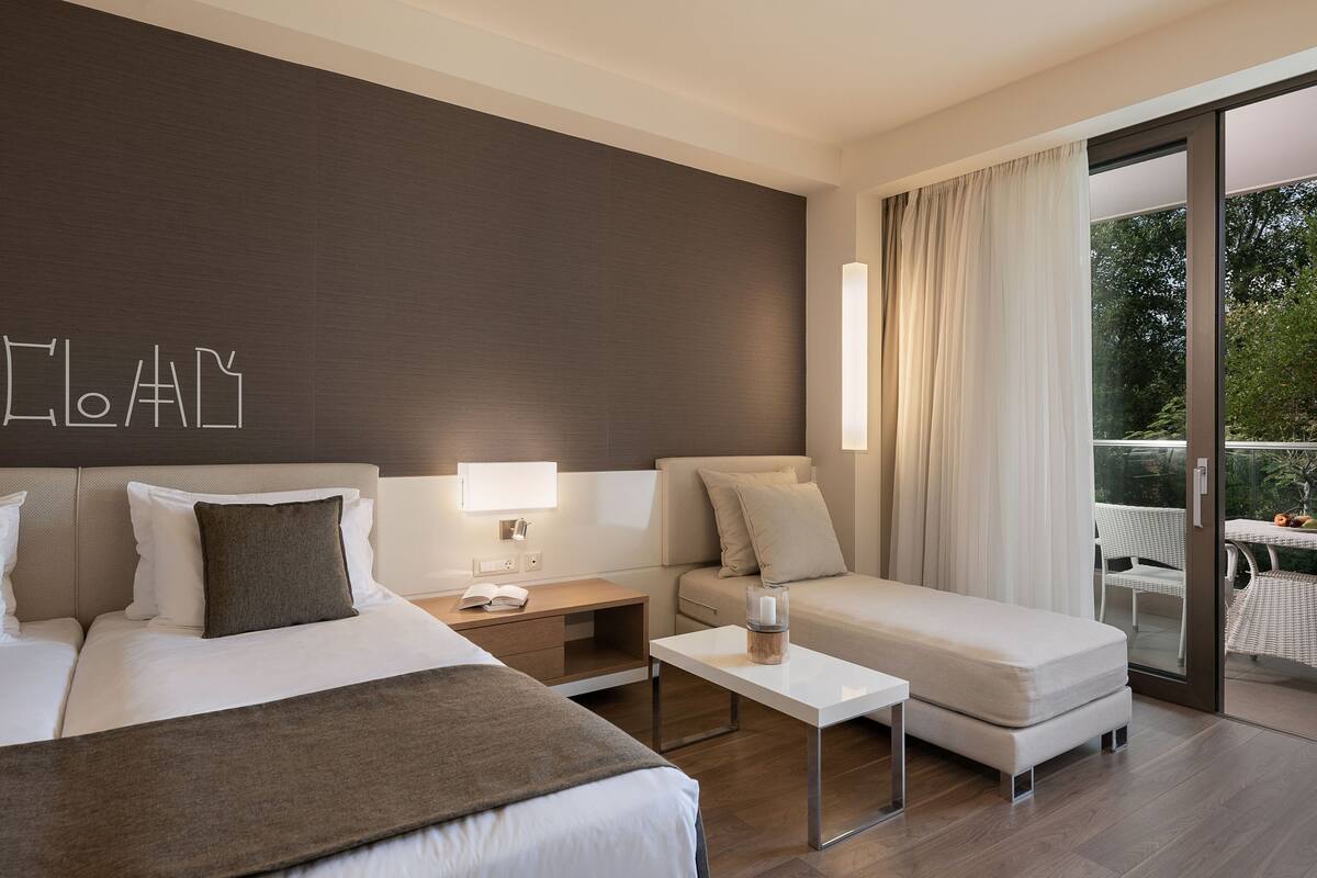 Una habitación de hotel moderna con una cama grande, un sofá, una mesita y un balcón con mobiliario de exterior.