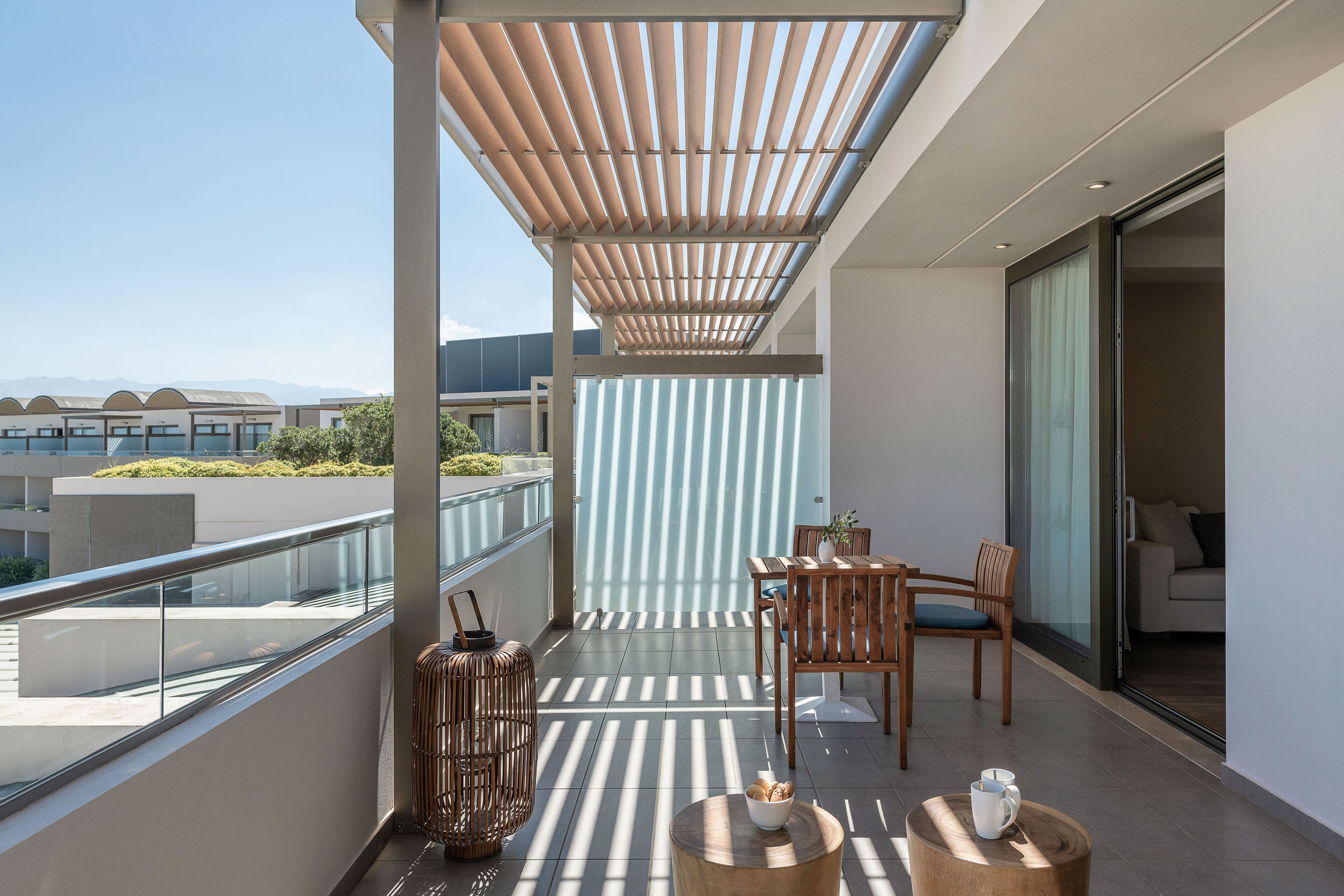 Un balcón con una pérgola de madera, barandilla de vidrio y zona de estar con mesa y sillas.