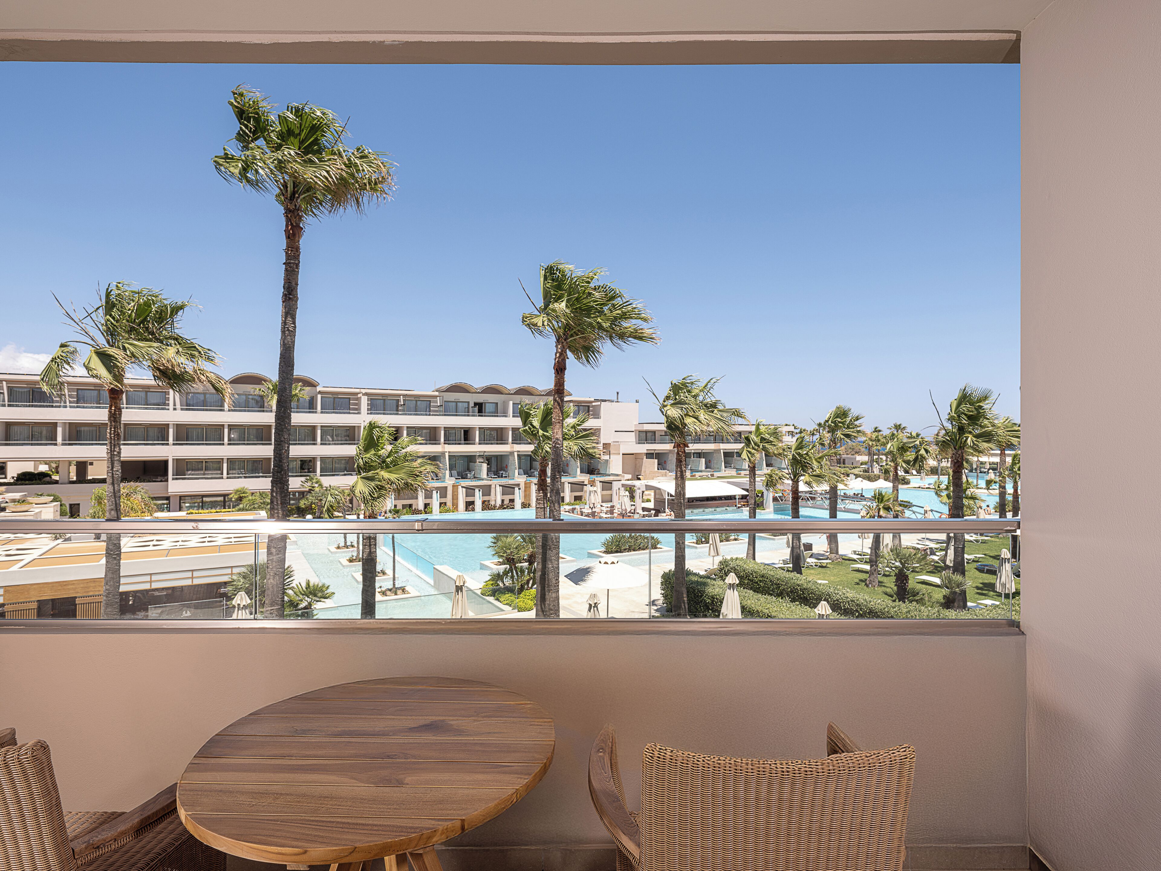 A balcony with a wooden table and wicker chairs, overlooking a pool area with palm trees and a hotel building in the background.