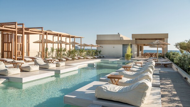 Zona de piscina con sillones de descanso y sombrillas, rodeada por un edificio moderno con vista despejada al mar.