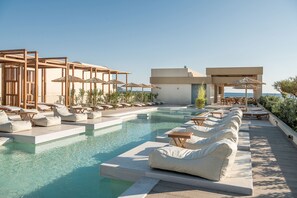 Zona de piscina con sillones de descanso y sombrillas, rodeada por un edificio moderno con vista despejada al mar.