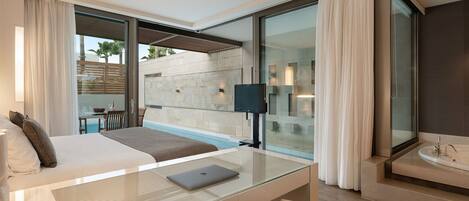 A modern hotel room with a glass desk, a bed, a laptop, a large window, and a bathroom with a bathtub.