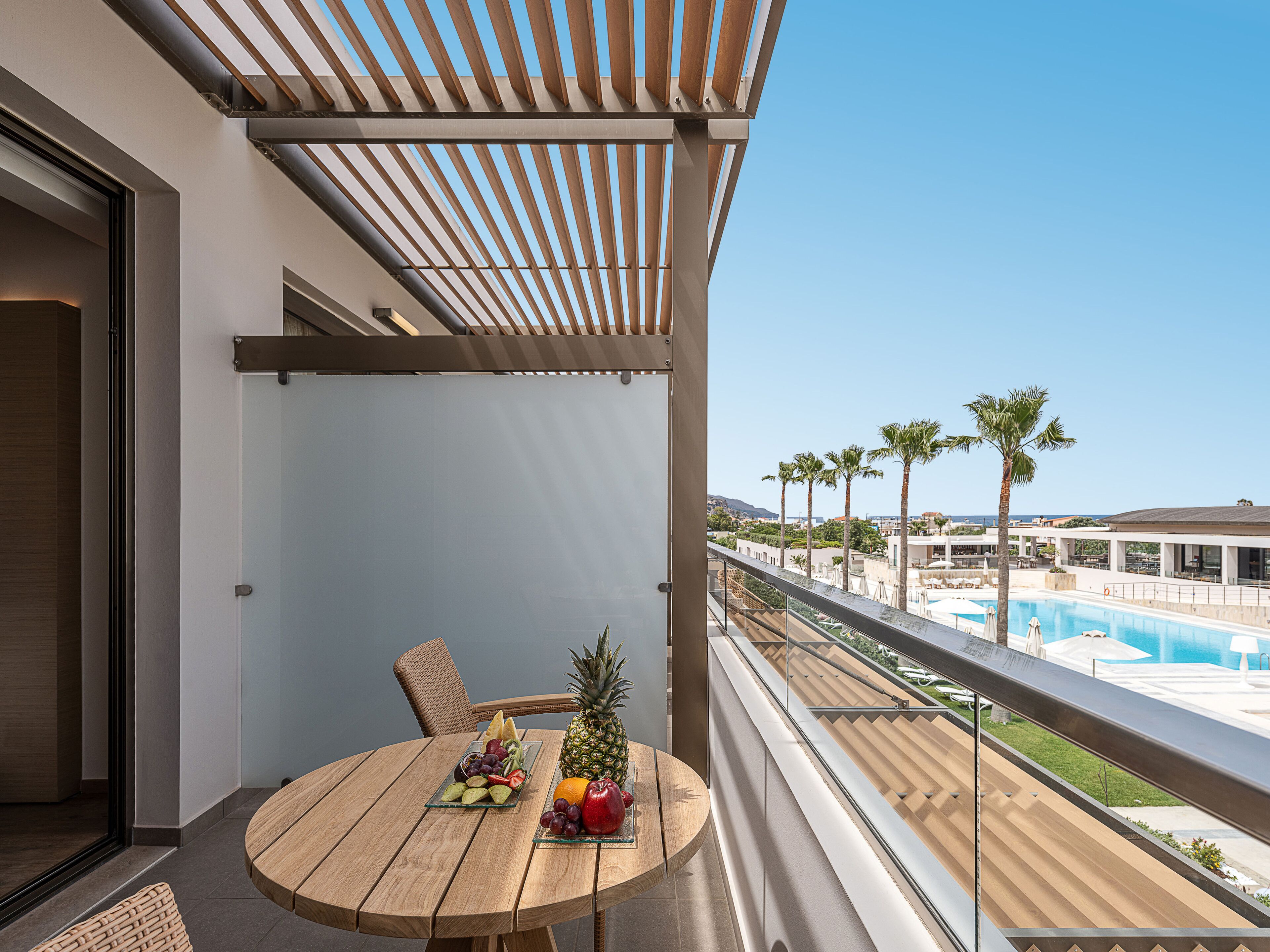 Un balcón con una mesa de madera dispuesta con fruta, con vistas a una piscina y palmeras.