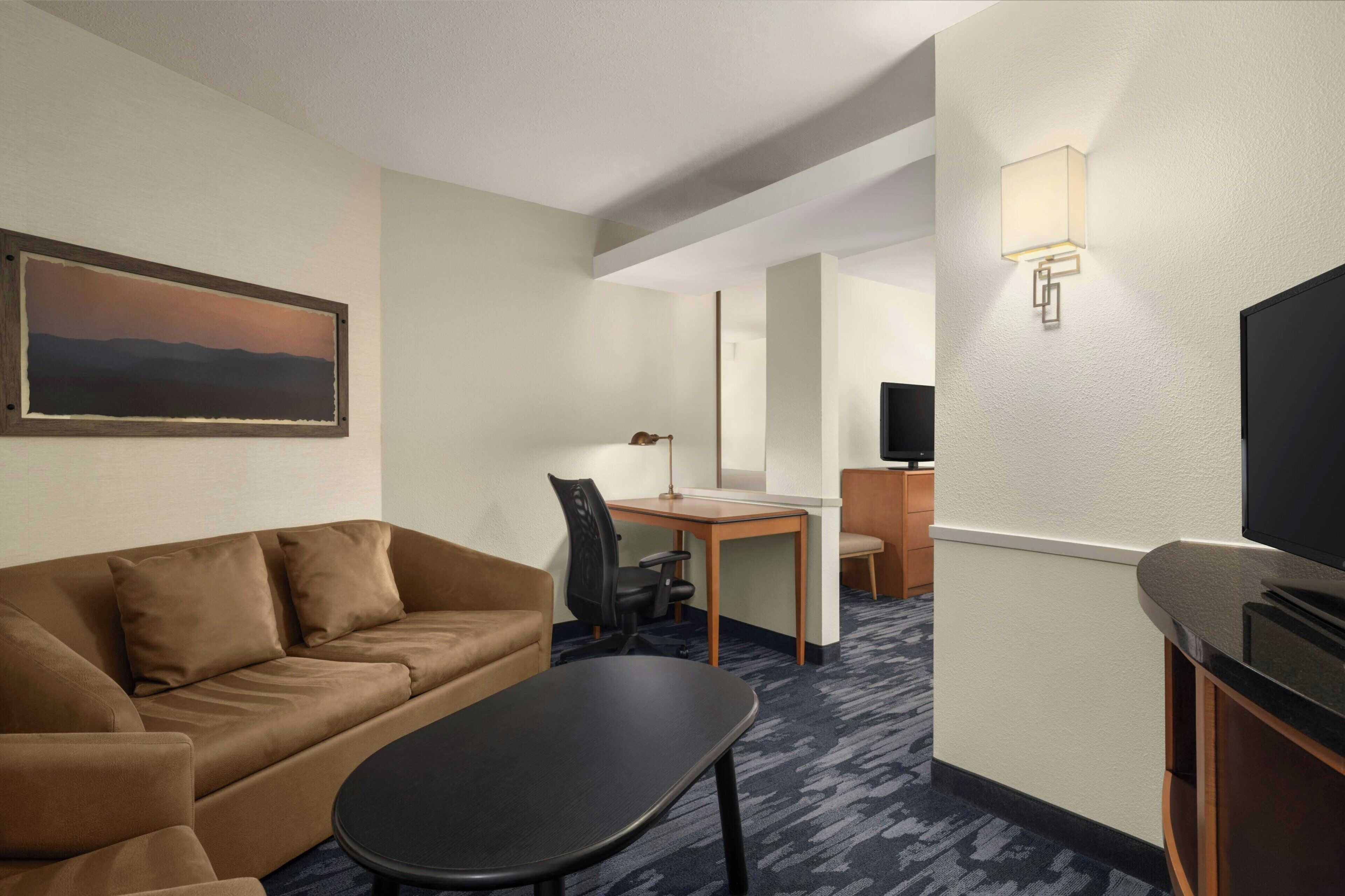 Habitación de hotel con sofá, escritorio, silla, televisión y un cuadro en la pared.