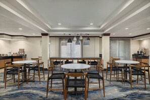 Un comedor de hotel con mesas y sillas redondas, una cocina al fondo y una alfombra estampada.
