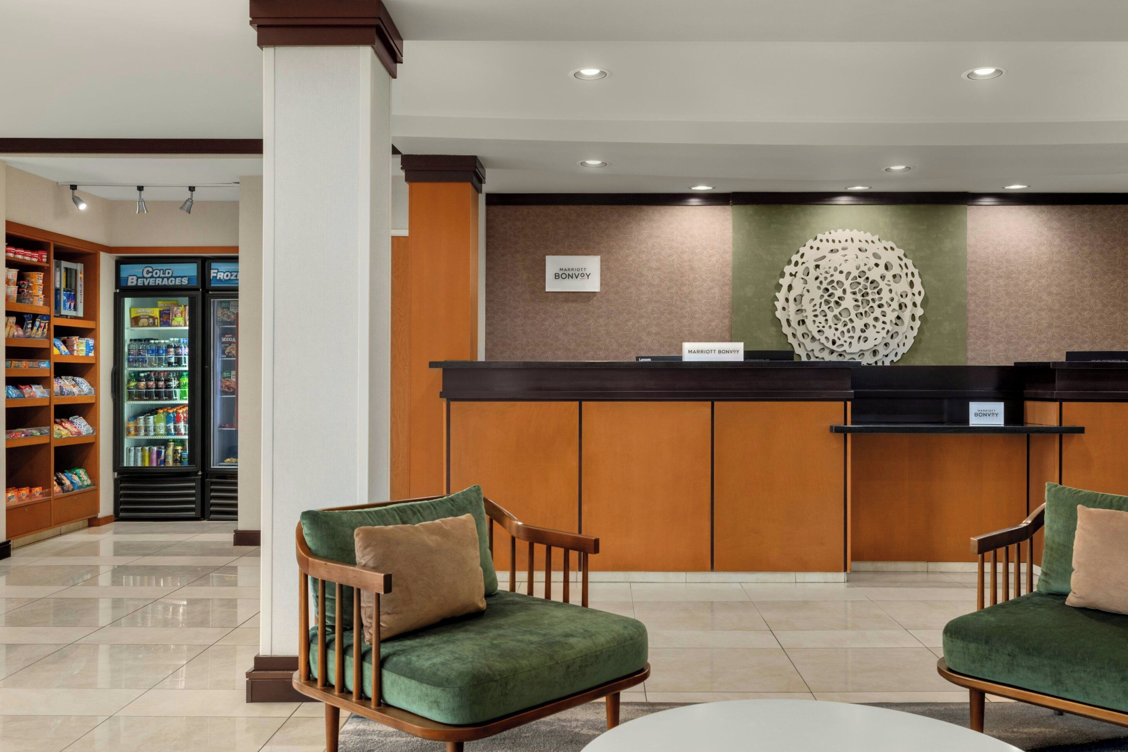 A hotel reception area with a counter, two green upholstered chairs, a vending machine, and a decorative wall piece.