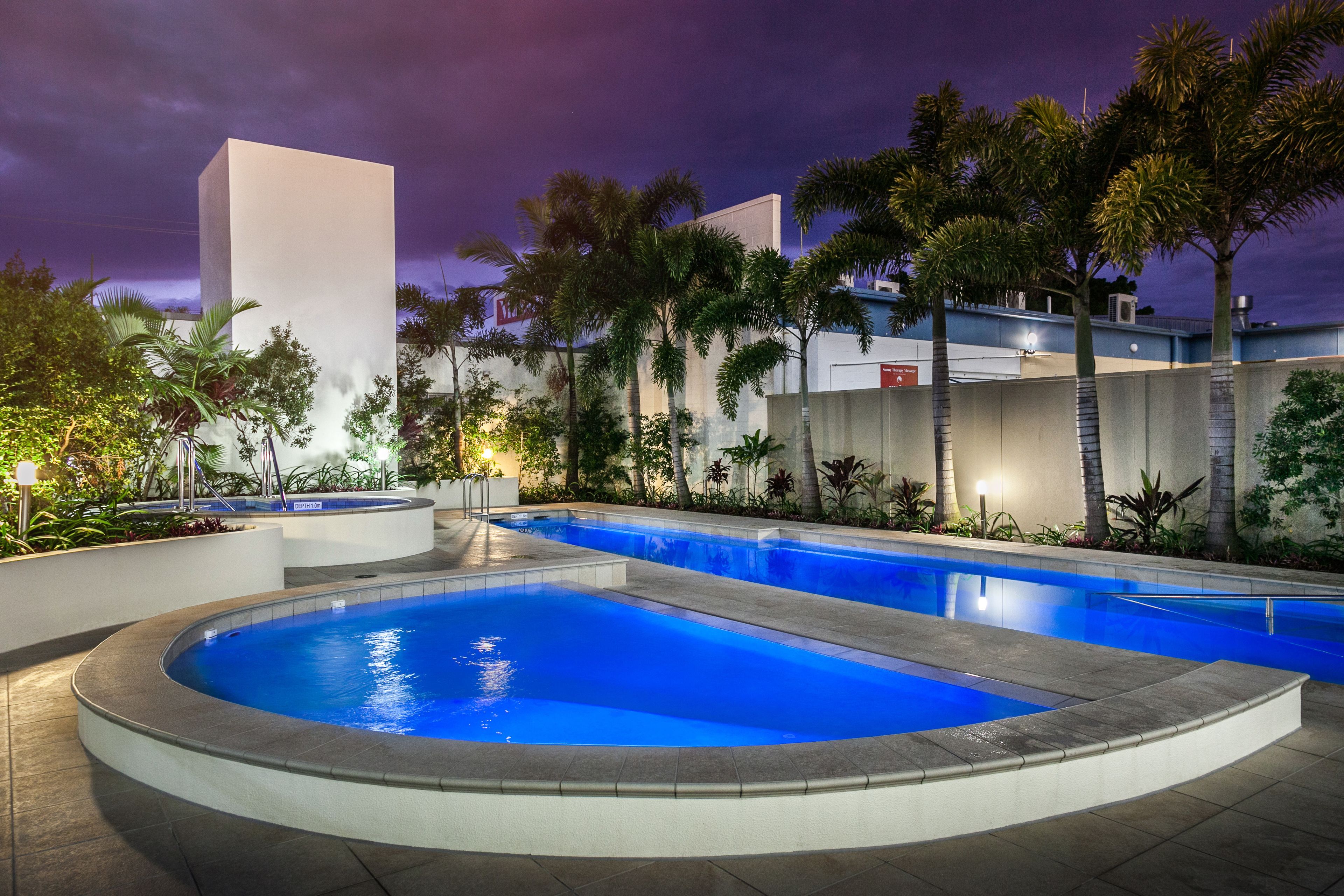 A swimming pool area with a curved section and a rectangular section, surrounded by palm trees and a building with a flat roof.
