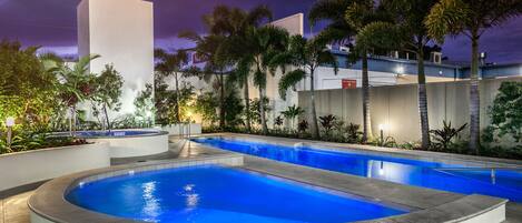 A swimming pool area with a curved section and a rectangular section, surrounded by palm trees and a building with a flat roof.