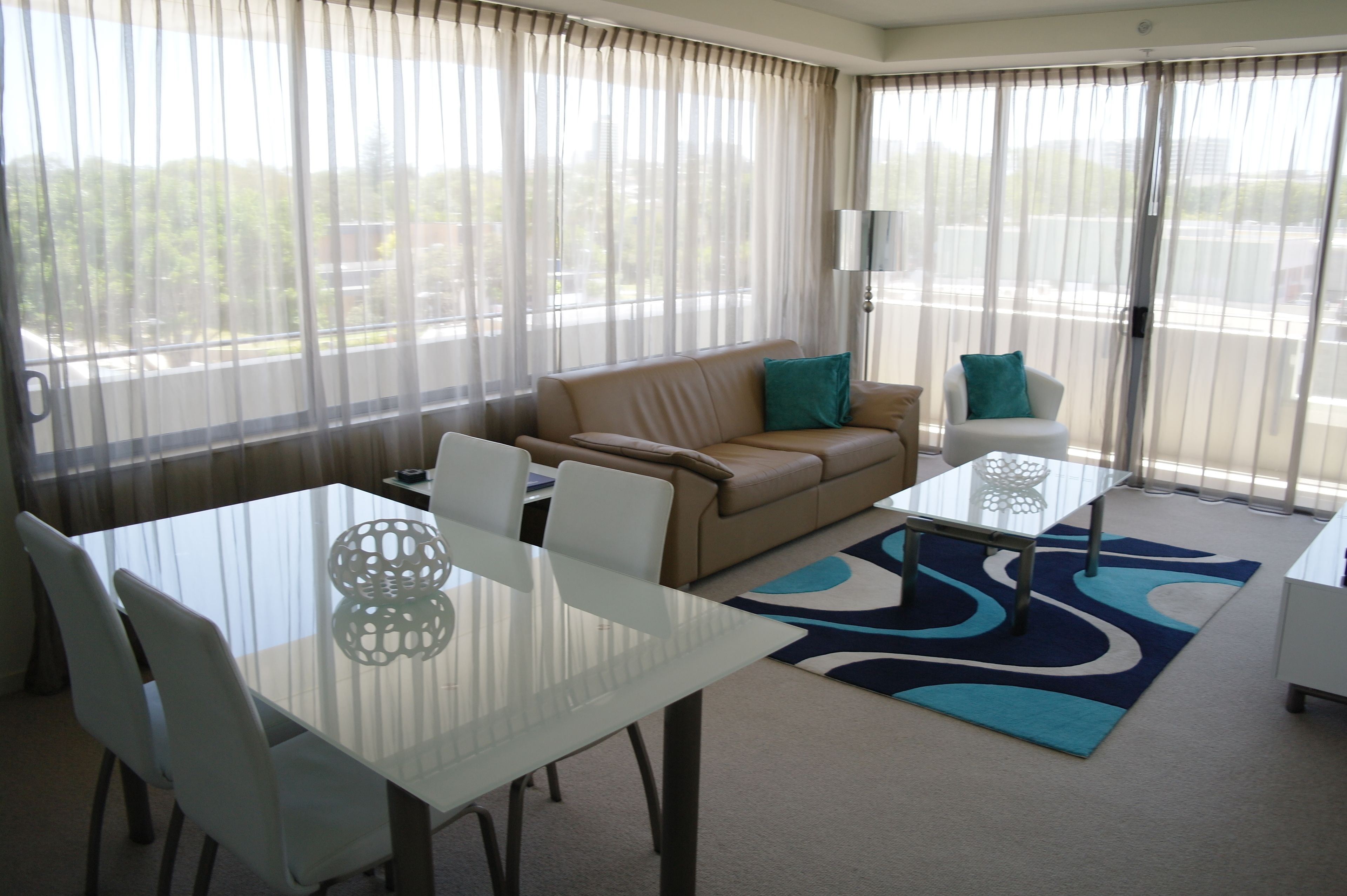 A modern hotel room with a glass dining table, beige sofa, and a small white side table.