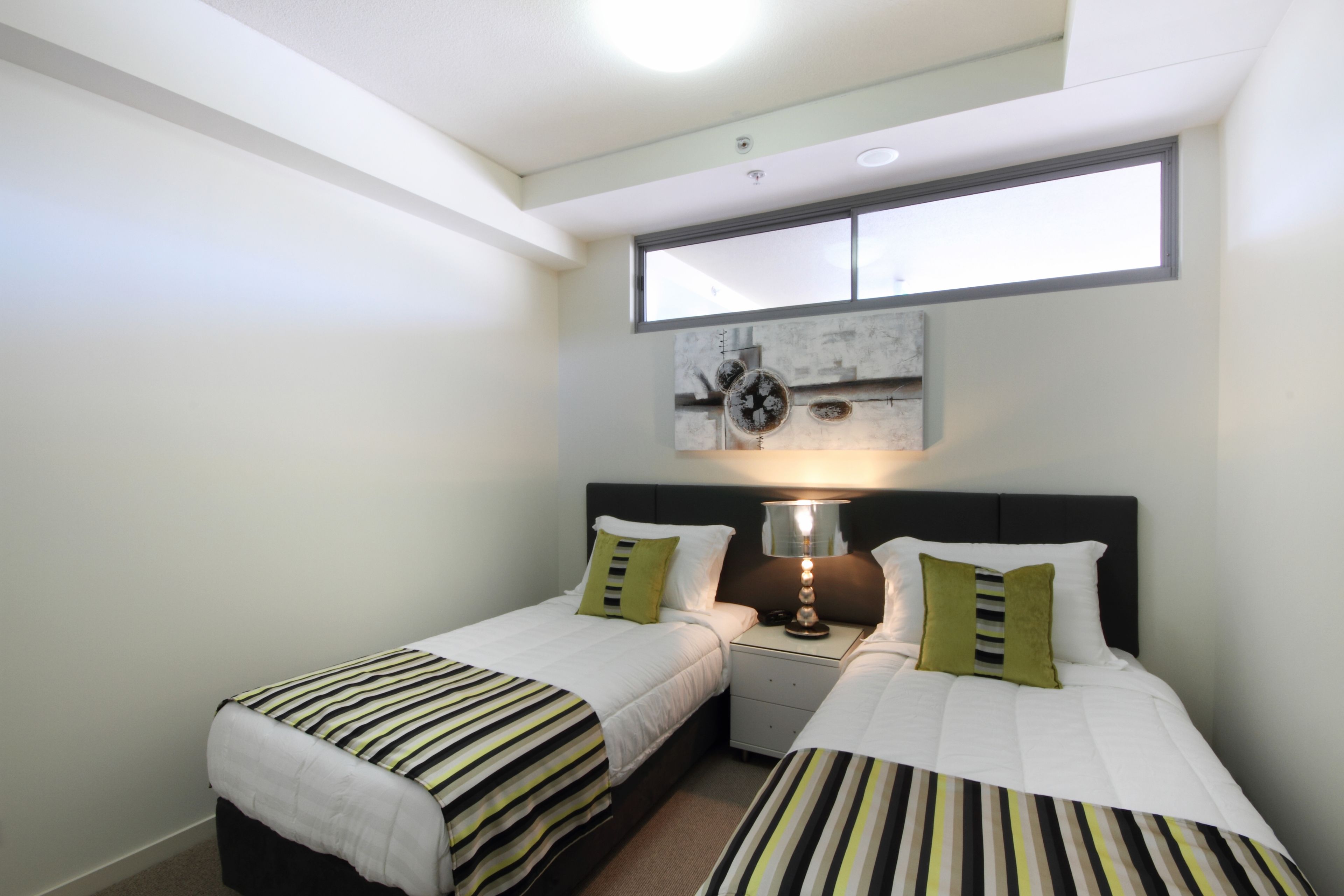Quarto duplo com duas camas, mesa de cabeceira, luminária e uma janela grande.