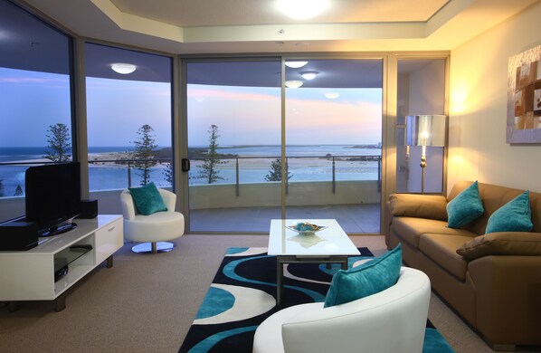 A modern living room with a large sliding glass door leading to a balcony with a view of the beach. - Monaco (Caloundra)