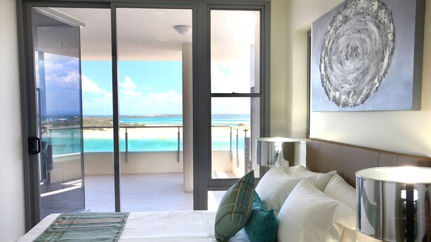 A hotel room with a bed, pillows, a lamp, and a view of the ocean through a sliding door.