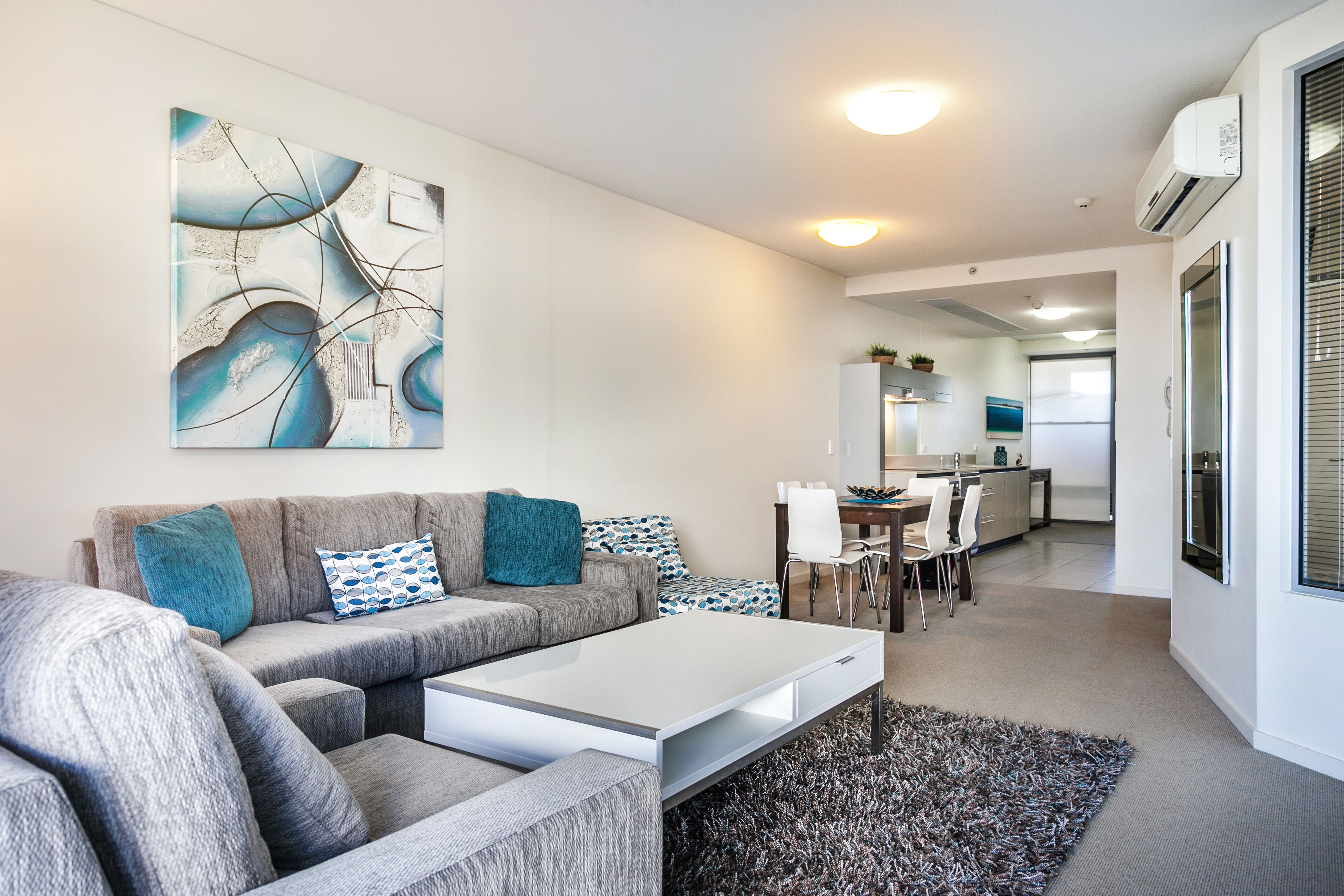 A modern living room with a sectional sofa, a coffee table, and a large abstract painting. There is a dining area with a table and chairs, and a kitchen in the background.